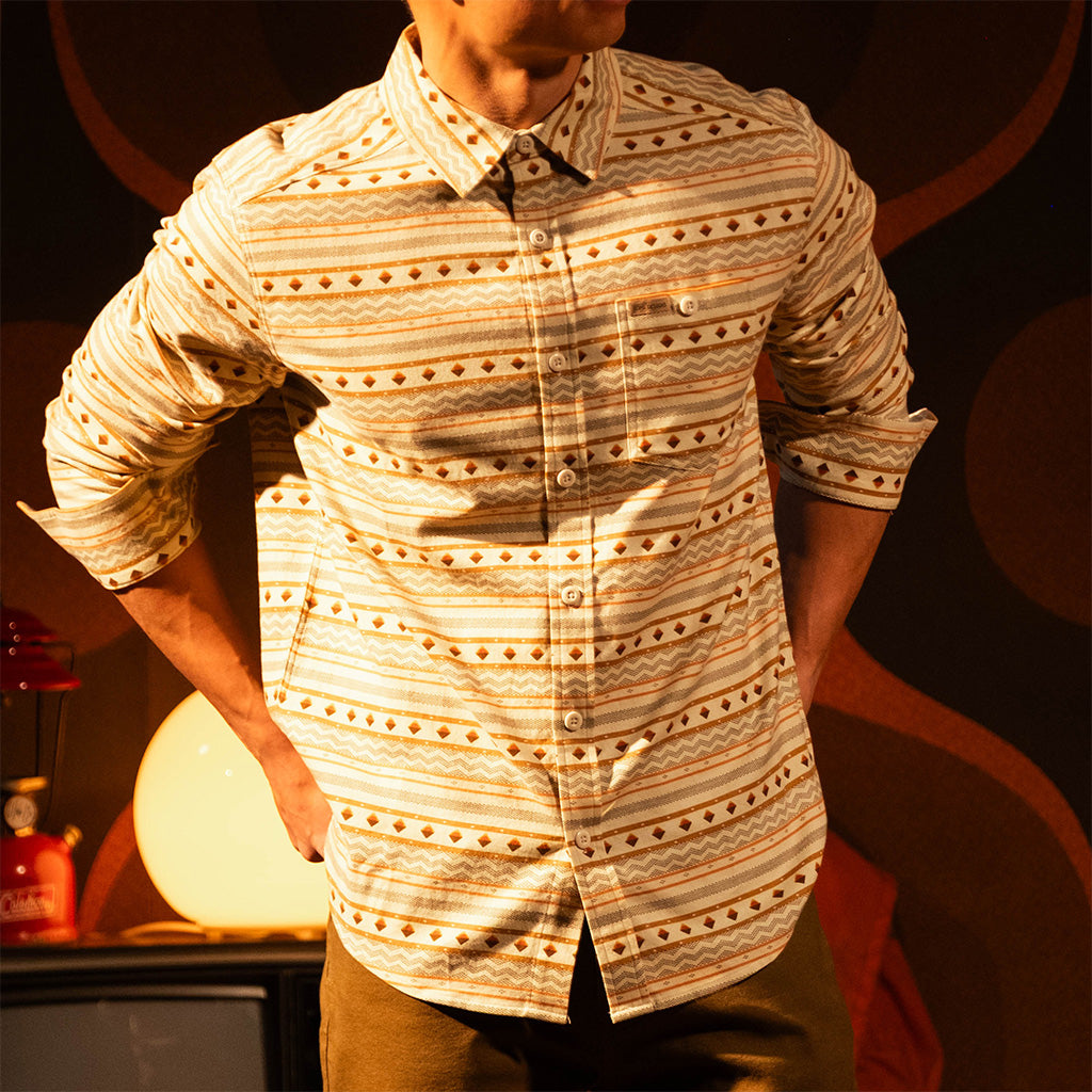 General lifestyle image of person wearing a "Bone White Sunset Stripe" Mountain Shirt Lightweight with a dark background with a warm glow in a dimly lit room. [Model is 5'11", wearing size medium]