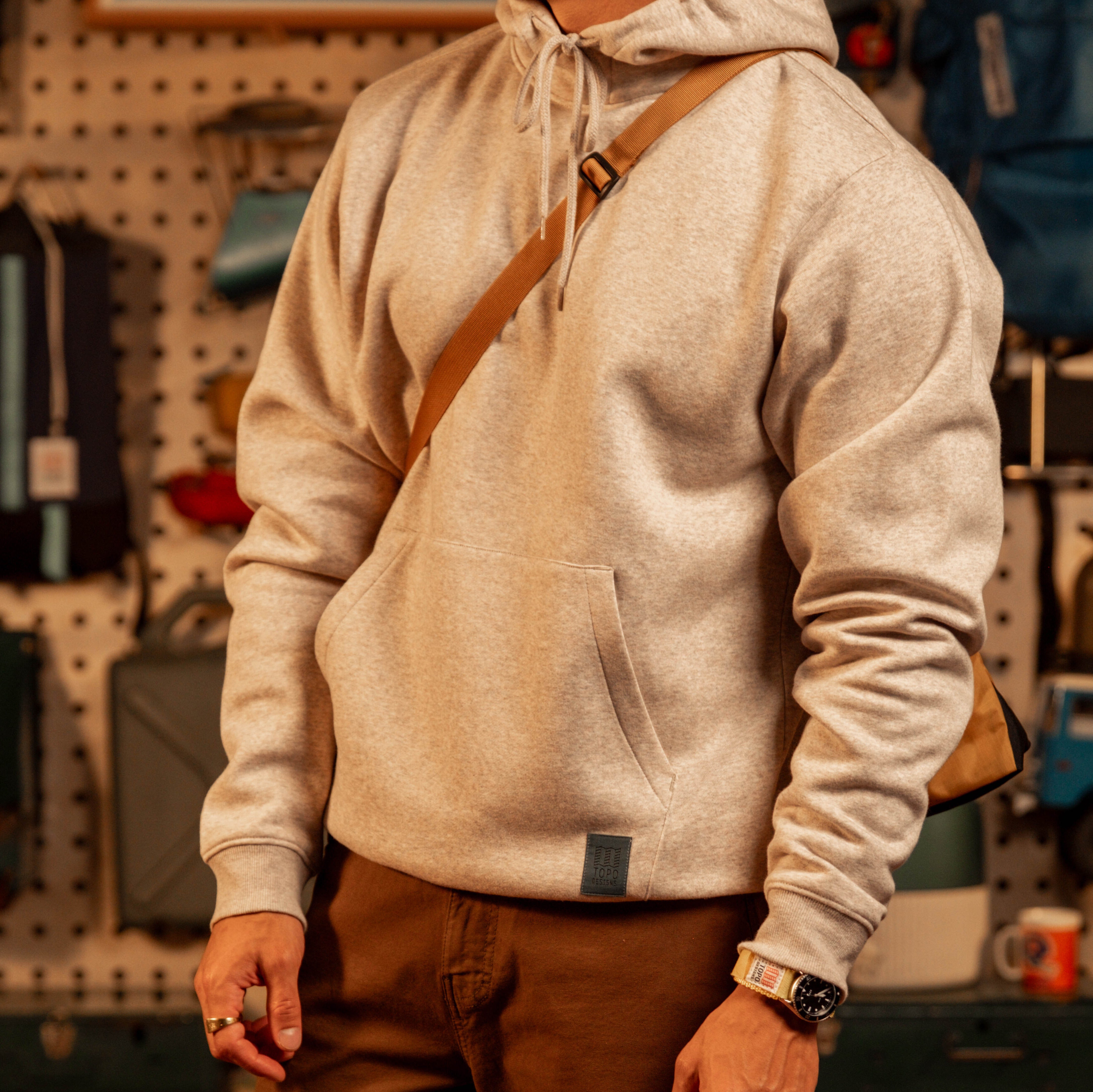 Person wearing a "Oatmeal Heather" hoodie with a brown strap over a pegboard wall. [Model is 5'11", wearing size medium]