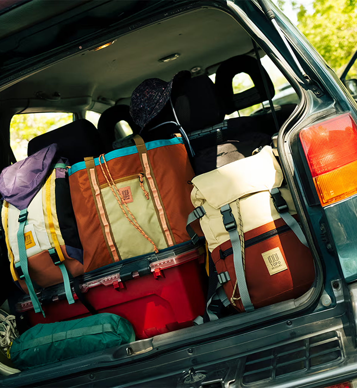 A close-up view of a vehicle's open trunk packed with camping gear, including a large brown and beige backpack, a red cooler, a cream roll-top bag, and other gear, with trees visible in the background.